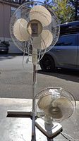 Large pedestal fan shown standing outdoors with one blade cracked, base is wide and rectangular.