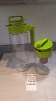 Tall clear plastic carafe with green handle and lid next to a smaller similar carafe with green lid and metal tea infuser inside