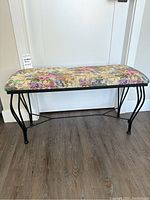 Front view of a rectangular bench with floral fabric upholstery on the seat and black wrought iron legs, placed on wood flooring against a white wall.