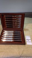 Open case showing six knives aligned in the lower half and six forks in the upper half, all with textured brown handles and shiny blades.