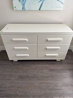 Front view of white 6 drawer dresser on wood floor with minor wear visible.
