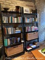 Two tall black metal frame bookshelves with wooden shelves and wicker baskets on lower shelves, filled with books and decorative items, placed against a brick wall.