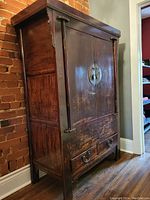 Closed view of Chinese wooden armoire showing dark wood, brass circular hardware on doors, brass ring pulls on two bottom drawers, wood patina, and decorative vertical wooden corner elements.
