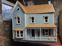 Front exterior view of blue two-story farmhouse dollhouse with white trim, bay window, wood shingle roof and front porch