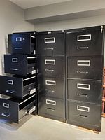 Three black metal filing cabinets placed side by side, all with four drawers partially open showing interior drawer space. Cabinets show minor surface wear and rust at the base. Two cabinets have security keys in locks, one cabinet is missing key.