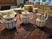 All three fine china serving pieces (teapot, sugar bowl with lid, creamer pitcher) on a colorful woven textile surface with other dishes in the background. White with gold stripes, elegant shape.