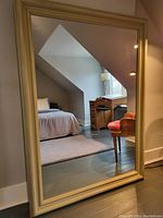 Full view of the wood framed mirror standing against a wall in a room with bed and desk reflected.