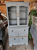 Front view of the grey painted wooden cabinet showing double-door open shelves on top, single drawer in the middle with metal ring pulls, and two closed doors with similar metal ring pulls at the bottom.