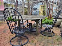 Photo of four black iron swivel patio chairs surrounding a rustic wooden outdoor table on a brick patio, showing the overall set and condition.