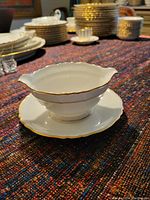 Gravy bowl with attached saucer, white with gold edging, shown on a textured fabric surface with additional china visible in background.