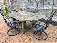 Side view showing the teak octagonal patio table with weathered grey surface and four black metal chairs arranged around it on a brick patio.