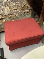 Red upholstered ottoman with no visible damage sitting on floor near stone wall.
