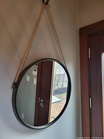 Round mirror hanging on a wall by a natural fiber rope. Black metal frame encircles the reflective glass. Reflections show a door and outdoor patio.