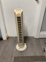 Front view of beige tower standing fan showing vertical vents and round base on wood floor.