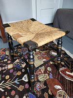 Front and top view of antique wooden stool with black painted legs and woven straw seat, displaying minor wear.