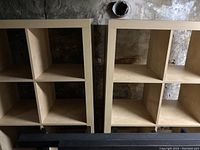 Two beige wooden shelving units, each with four large square compartments, shown standing vertically against a stone wall