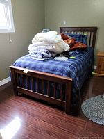 Front angle of wooden single bed frame with headboard and footboard, placed in bedroom with some bedding and household items on top of mattress not included in sale