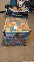 Front view of square storage box with colorful geometric pattern and metal reinforced edges and corners, sitting on carpeted floor.