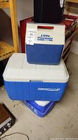 Three stacked coolers. Bottom is white and blue Coleman Polylite 40 cooler, middle is a blue and white Igloo Little Playmate cooler with red handle.