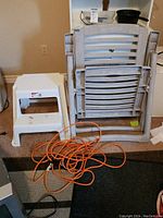 Four white plastic folding lawn chairs stacked together with an orange extension cord coiled in front, and two white plastic step stools placed nearby on carpeted floor.