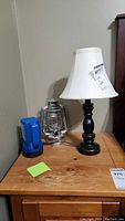 Table lamp with white shade, blue flashlight, and silver lantern on wooden surface against a wall.