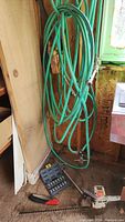 Green garden hose hanging on the wall with metal connectors, drill bit set, garden saw with black handle on the floor, and Black and Decker hedge trimmer partially visible.