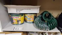 Two 75 ft green expandable Pocket Hoses in original packaging and a coiled green hose on a shelf with floral coffee cups below