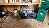 Overview of the shed area showing the black plastic basket with paint rollers and trays, tire leaning against the wall, paint can, and hand tools below the workbench.
