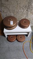 Four brown metal weight plates placed on white bench and floor, set of two 25 lbs and two 10 lbs.