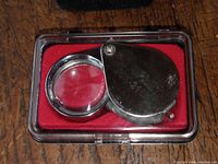 Jeweler's loupe folded closed inside clear plastic case with red velvet-like interior, visible metal housing and lens, photographed on wooden surface.