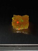 Rough fire opal specimen with intense orange color, green and red play-of-color flashes on dark background.