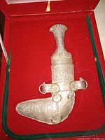 Gurkha kukri fully sheathed in silver scabbard displayed on red velvet with green trim inside display box.