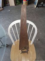 Full length view of the antique hand saw resting on a white chair, showing wooden handle and rusty blade