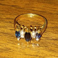Close-up photo of yellow gold ring with three marquise-cut dark blue sapphires and four small diamonds on wood surface.