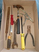 Photo showing nine old hand tools inside a cardboard box: claw hammer, pliers, 2 screwdrivers, wrench, file/saw blade, locking plier, utility knife, and awl tool.
