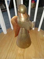Front view of brass and copper angel ornament showing full figure holding a book with copper wings, placed on wooden surface near white railing.