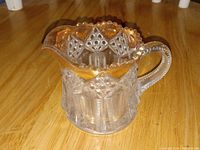 Clear pressed glass cream pitcher with intricate embossed diamond and geometric patterns, gilt gold scalloped rim, and curved handle.