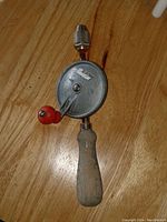 Front view of the hand auger drill showing wooden handle, circular metal gear with crank handle, and metal chuck.