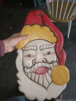 Front close-up photo of hand carved wooden Santa Claus mask showing red hat with yellow trim and pom pom, white beard, and detailed facial features.