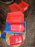 Three plastic storage bins in red and blue shown stacked. Bins have compartments for sorting small items like drill bits, nails, nuts, and bolts. Varying sizes and colors visible.