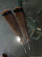 Pair of long hawk feathers laid side by side on a glass table, showing the brown color with black tops and patterned markings along the shafts.
