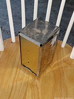 Napkin holder positioned on a wood chair showing top and one side view, displaying stainless steel material with visible surface marks and corner notch details.