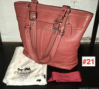 Photo showing front of pink textured leather Coach handbag with double buckled shoulder straps, leather Coach tag, and dust bag and fabric strap placed beside it.