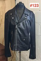 Full front view of black leather biker jacket displayed on mannequin against white wall.