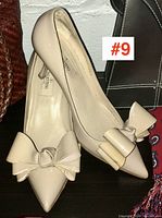 Pair of ivory Valentino ladies pointed toe heels with large bow detail, showing side and top angles on dark surface with handbags in background.