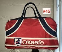 Oblique front view showing red front panel with navy and white stripes, black handles, O'Keefe logo patch, and cursive writing below logo.