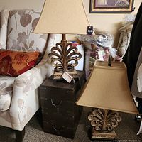 View of two aged bronze decorative table lamps with matching beige fabric shades, placed side by side on a small dark wooden chest next to a chair.