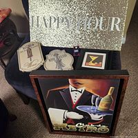 Overview photo showing most of the festive bar signs stacked together, including a glittery 'Happy Hour' sign, small wooden bar themed plaques, and a large framed illustration of a waiter holding wine and glasses.