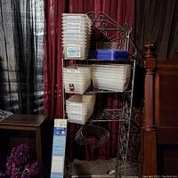 Full view of metal bathroom tower with several plastic storage boxes and a box of window blinds beside it, showing overall items in the lot.