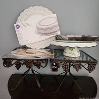 Photo showing two metal cake display stands, ceramic cake stand with leaf pattern, Wilton cake cards and doilies, gold-tone cake server